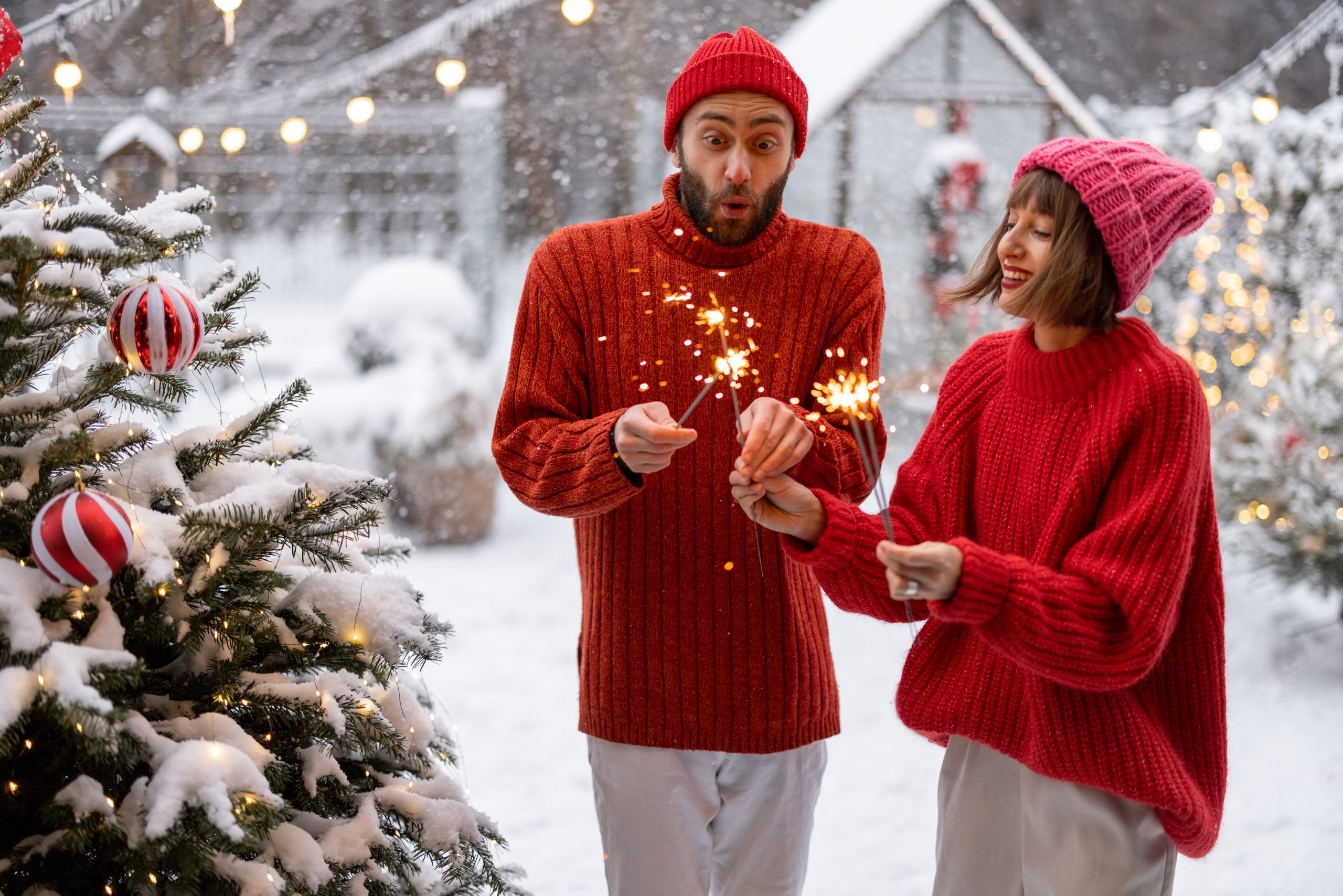 Man and woman in red sweaters celebrate New Year's holidays by lighting sparklers and having fun near Christmas tree at snowy backyard